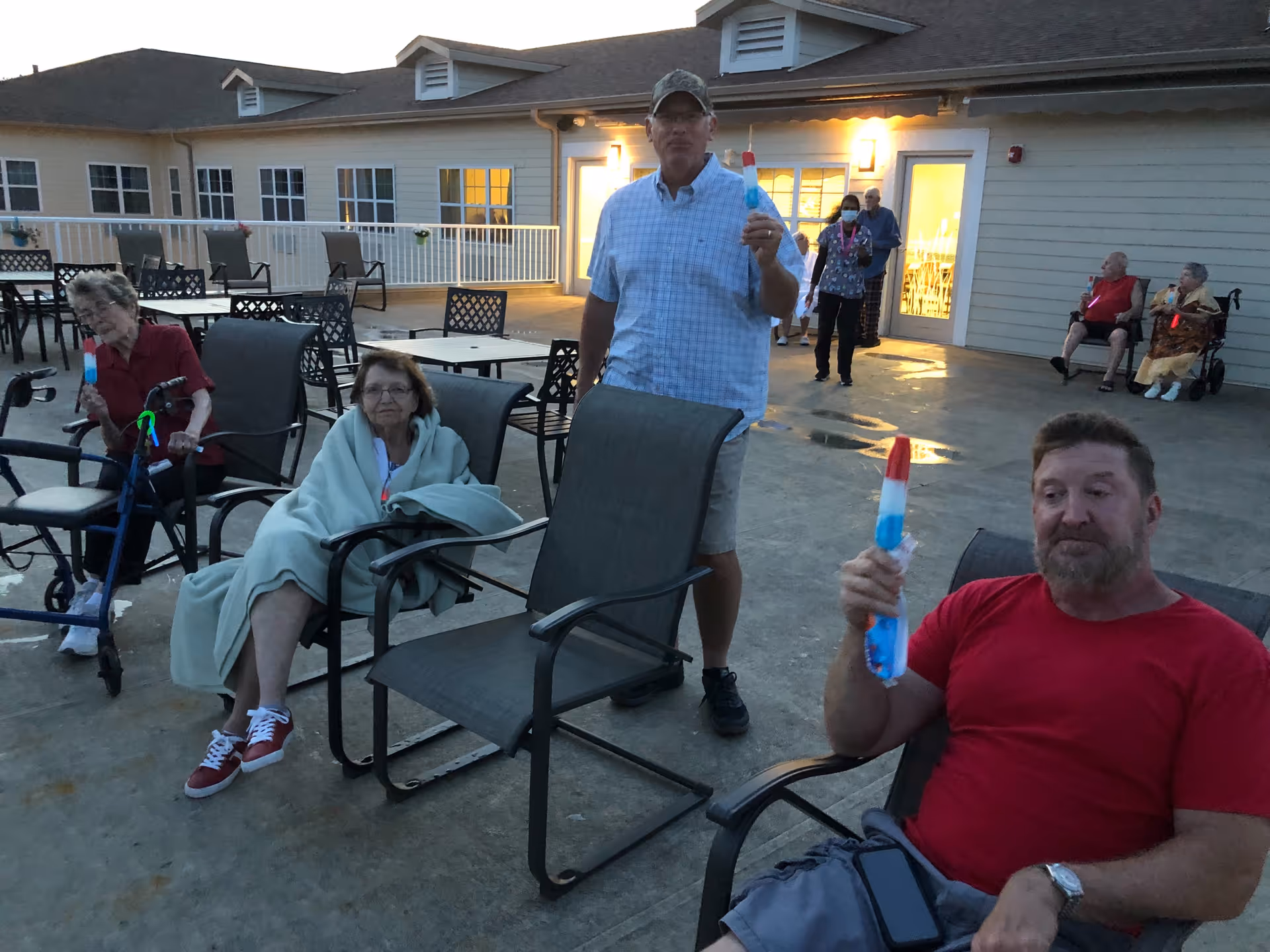 A group of elderly people and two men are outside on a patio in the evening. Some are seated in chairs, one woman is wrapped in a blanket, and several people are holding red, white, and blue popsicles. The building behind them has lit windows and doors.