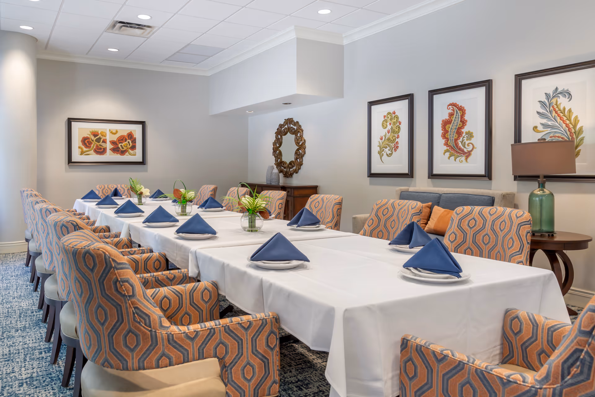 A dining room with a long table covered with a white tablecloth, set with plates and blue folded napkins. The room features patterned upholstered chairs around the table, floral artwork on the walls, a decorative mirror, and a lamp on a side table.