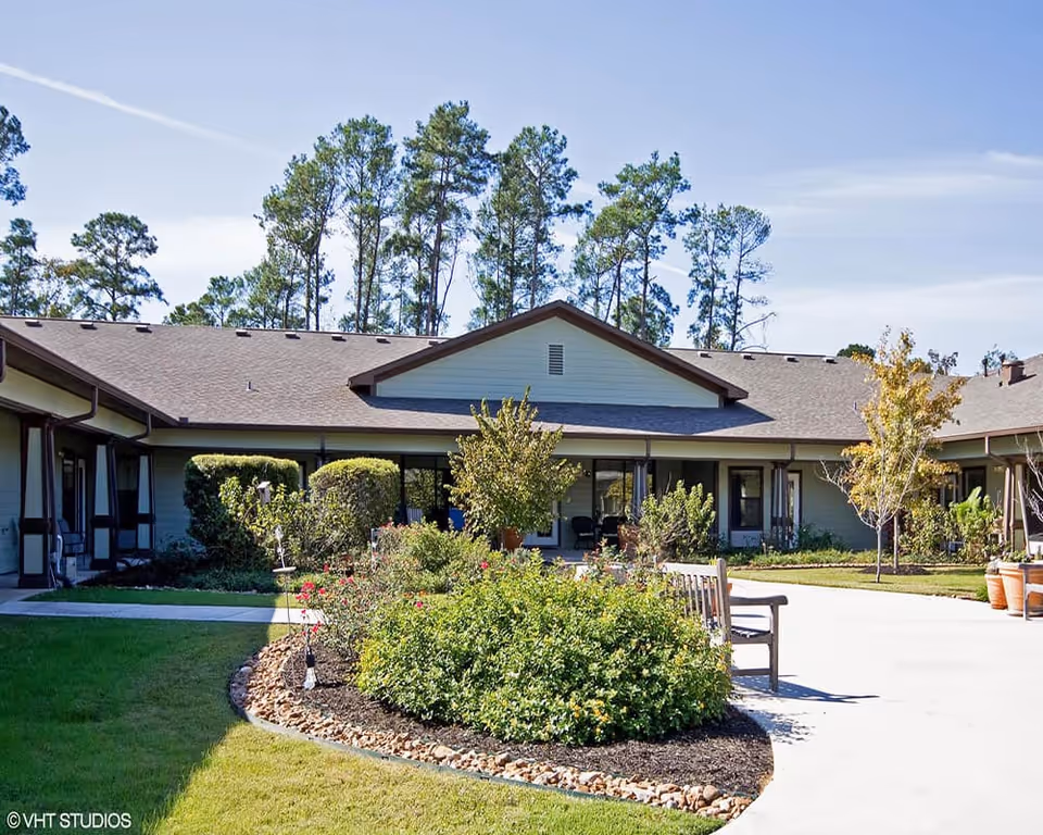 Outdoor courtyard area of Keepsake Village of Columbus Memory Care featuring a garden with shrubs and flowers, a wooden bench, and a single-story building with large windows and a pitched roof surrounded by tall pine trees under a clear blue sky.