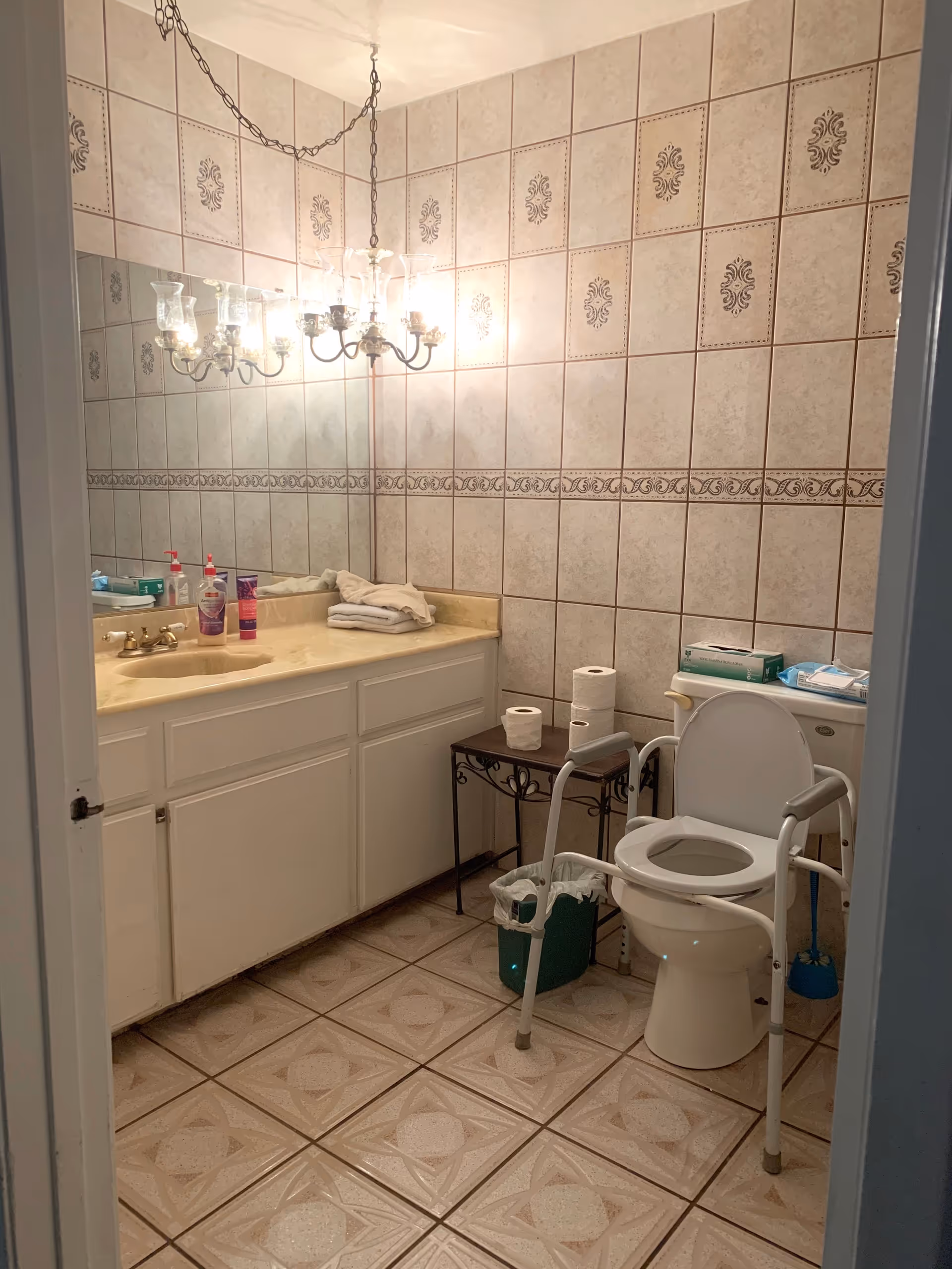 A bathroom with beige patterned tiles on the walls and floor. There is a white vanity with a beige countertop, a large mirror above it, and a chandelier-style light fixture. On the countertop are hand soap, lotion, and folded towels. Next to the vanity is a small table holding three rolls of toilet paper. The toilet has a white frame support rail around it, and there is a green trash bin nearby.