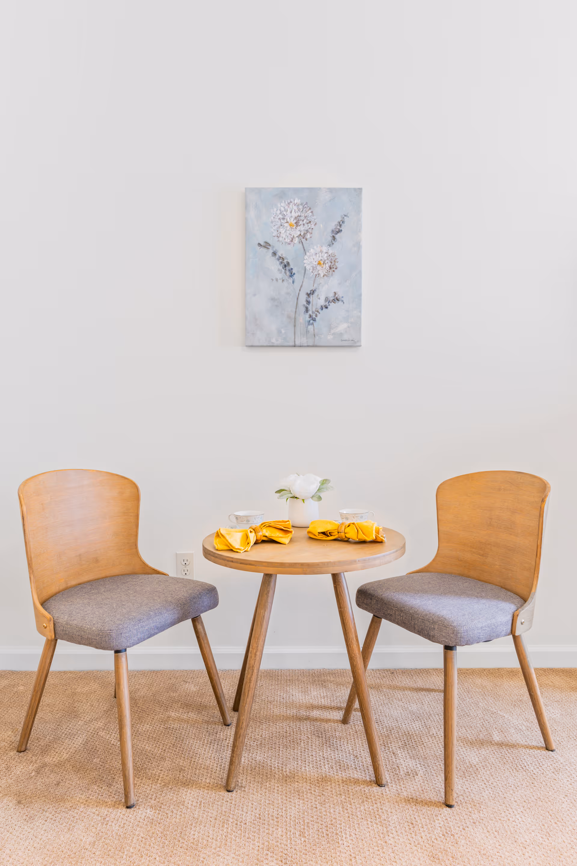 Small dining nook with a round wooden table set for two, two chairs, and a floral painting on a white wall.