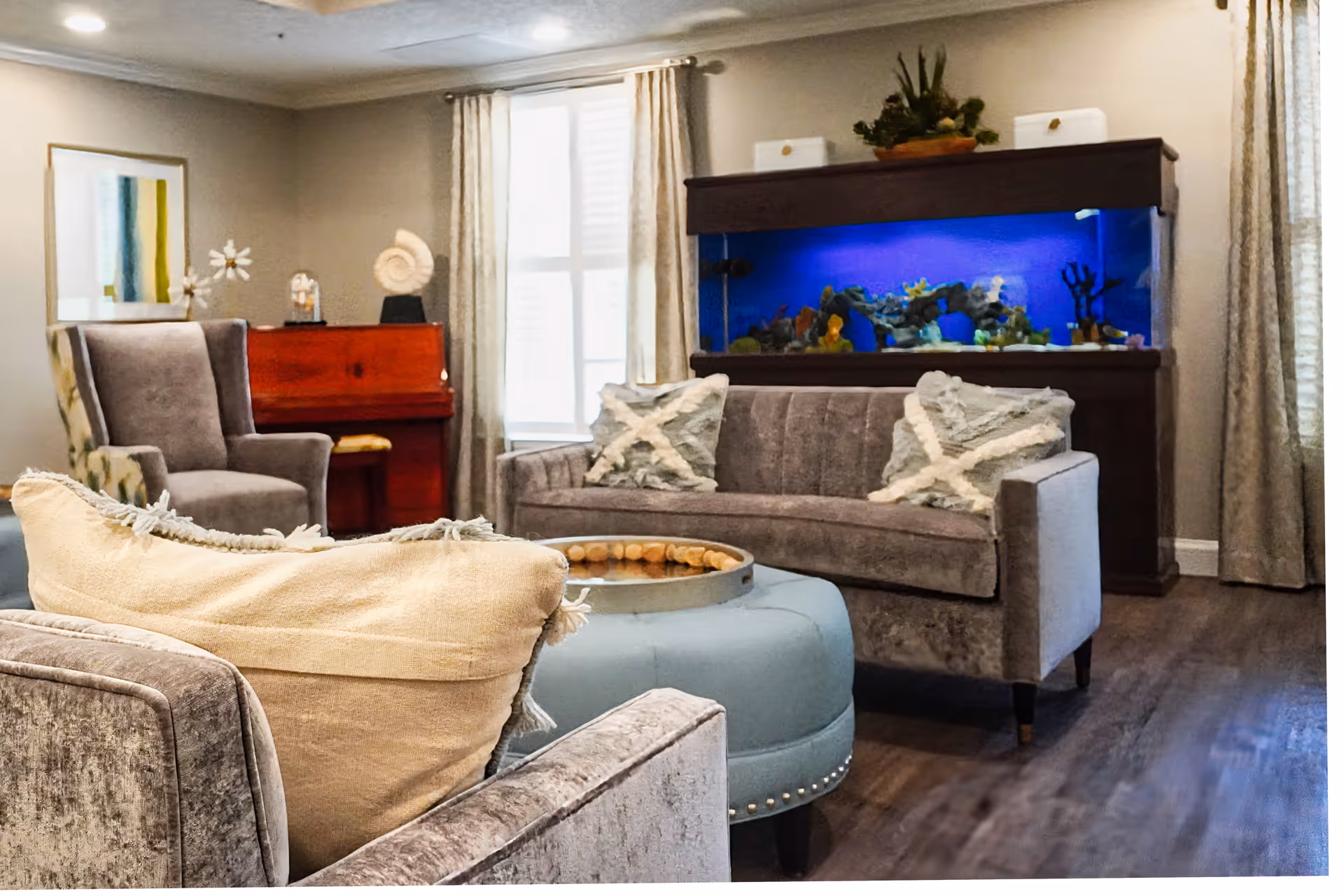 A cozy living room with a gray sofa adorned with two decorative pillows, a matching armchair with a beige pillow, and a round blue ottoman with a tray on top. In the background, there is a large aquarium with blue lighting, a wooden piano, and a window with light-colored curtains.