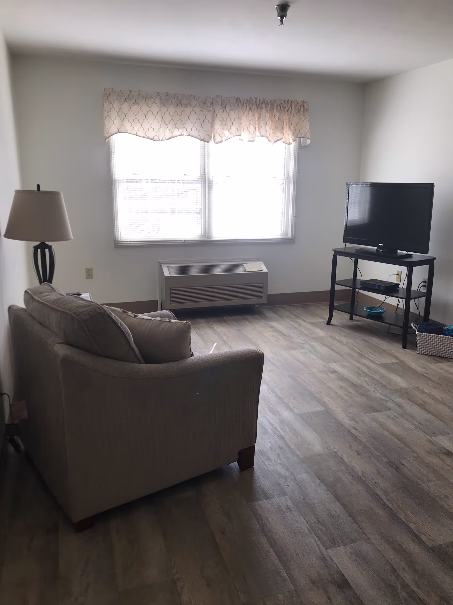 A small living room with a beige couch facing a flat-screen TV on a black stand. There is a floor lamp next to the couch and a large window with a patterned valance letting in natural light. The floor is wood-style laminate.