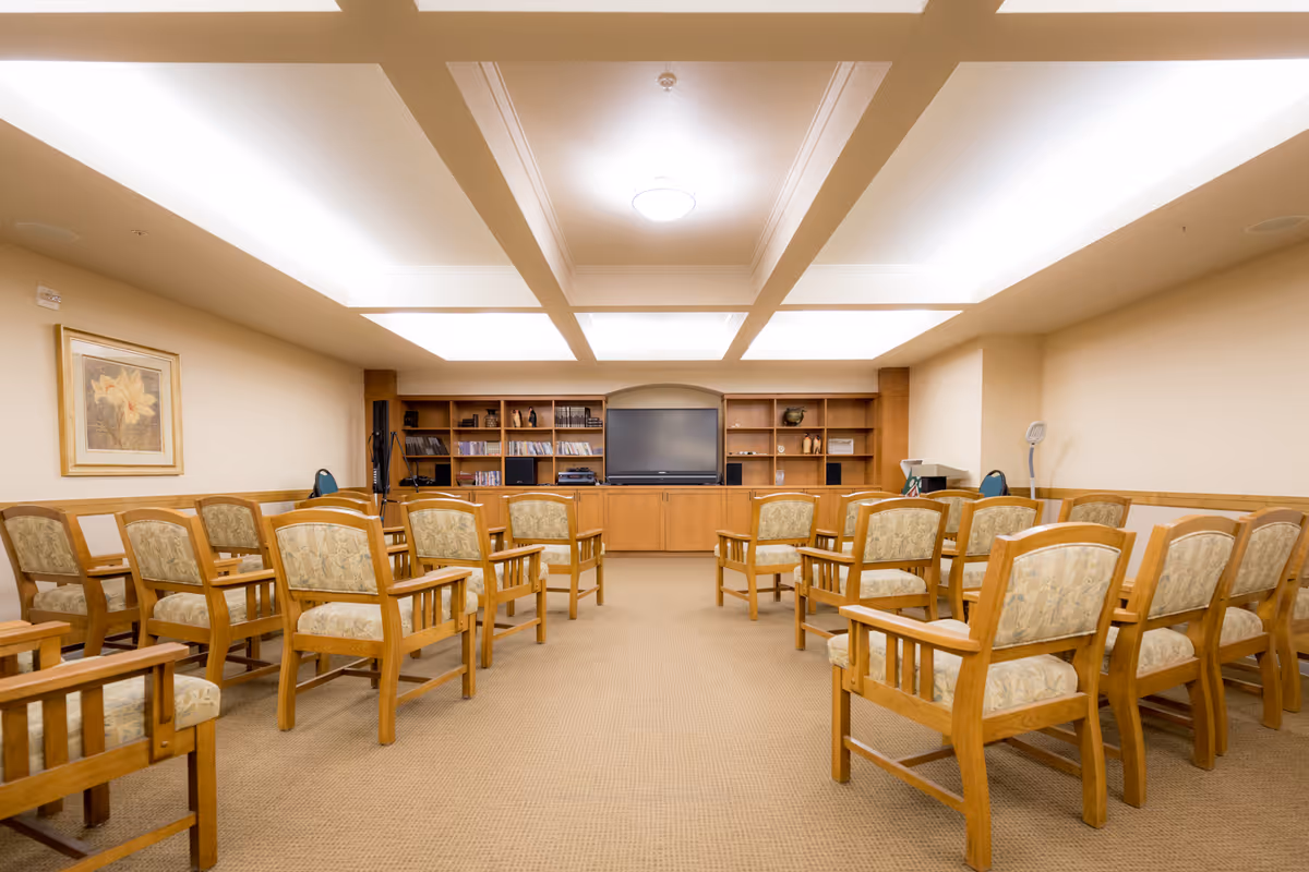 A well-lit room with rows of wooden chairs with patterned cushions facing a large flat-screen TV mounted on a wooden cabinet with shelves containing books and decorative items. The ceiling has recessed lighting panels, and there is a framed flower artwork on the left wall.