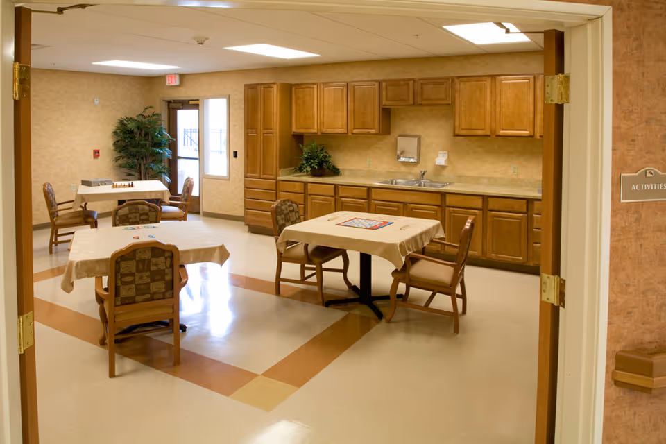 A well-lit activity room with tables and chairs arranged for games and socializing. The room features wooden cabinets and a countertop with a sink along one wall. There are board games on the tables, a potted plant in the corner, and a door leading outside. The room has beige walls and a patterned floor.
