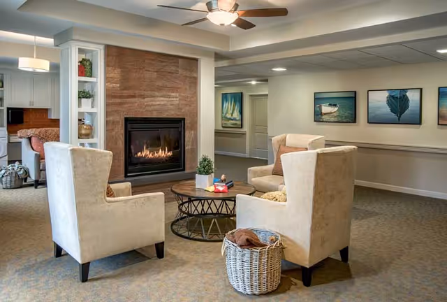 Cozy common lounge with four armchairs arranged around a round coffee table facing a lit fireplace, with artwork on the walls and a ceiling fan.