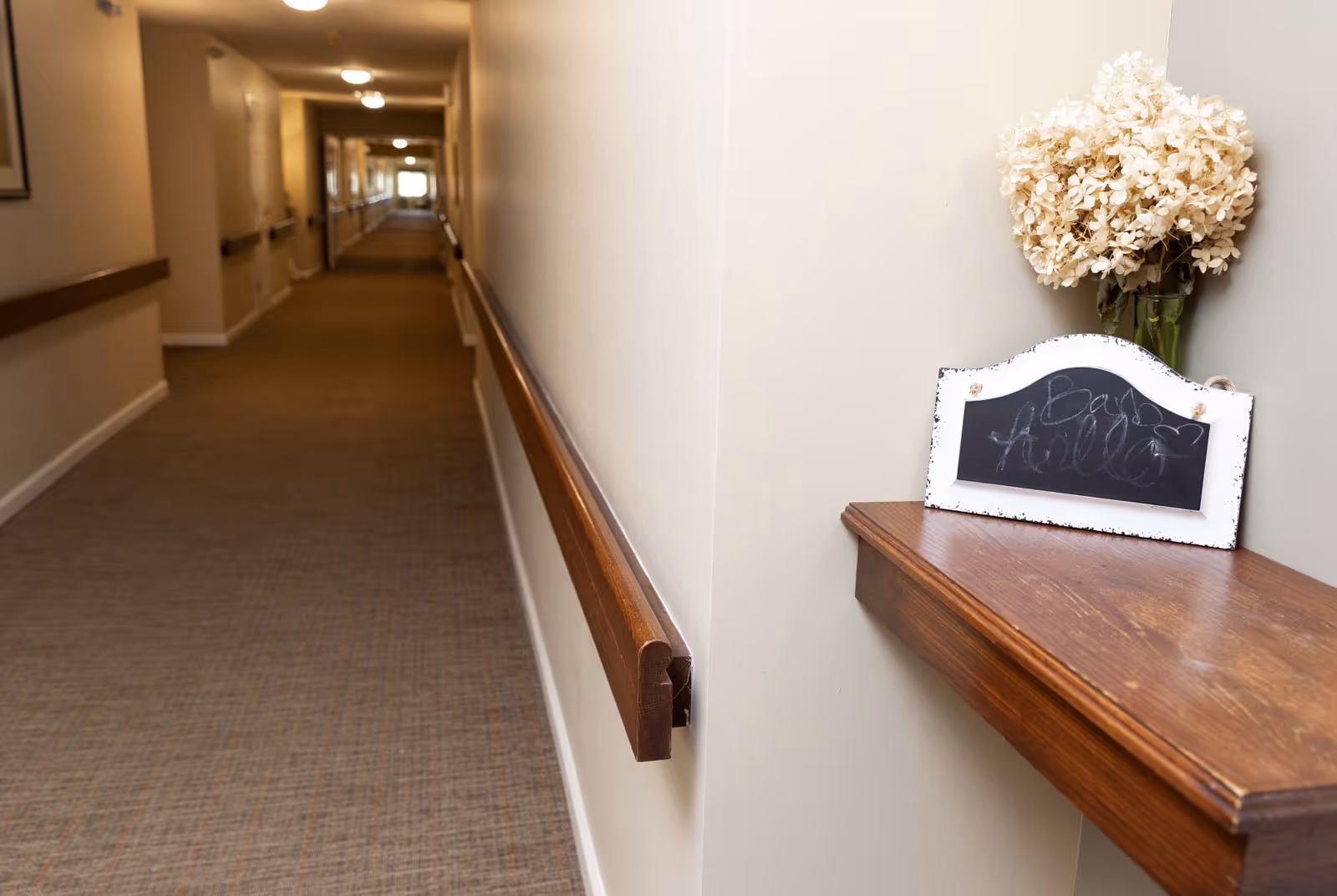 A long, carpeted hallway in a senior living facility with beige walls and wooden handrails on both sides. On the right side, there is a small wooden shelf holding a vase with dried white flowers and a small chalkboard sign with faint writing.