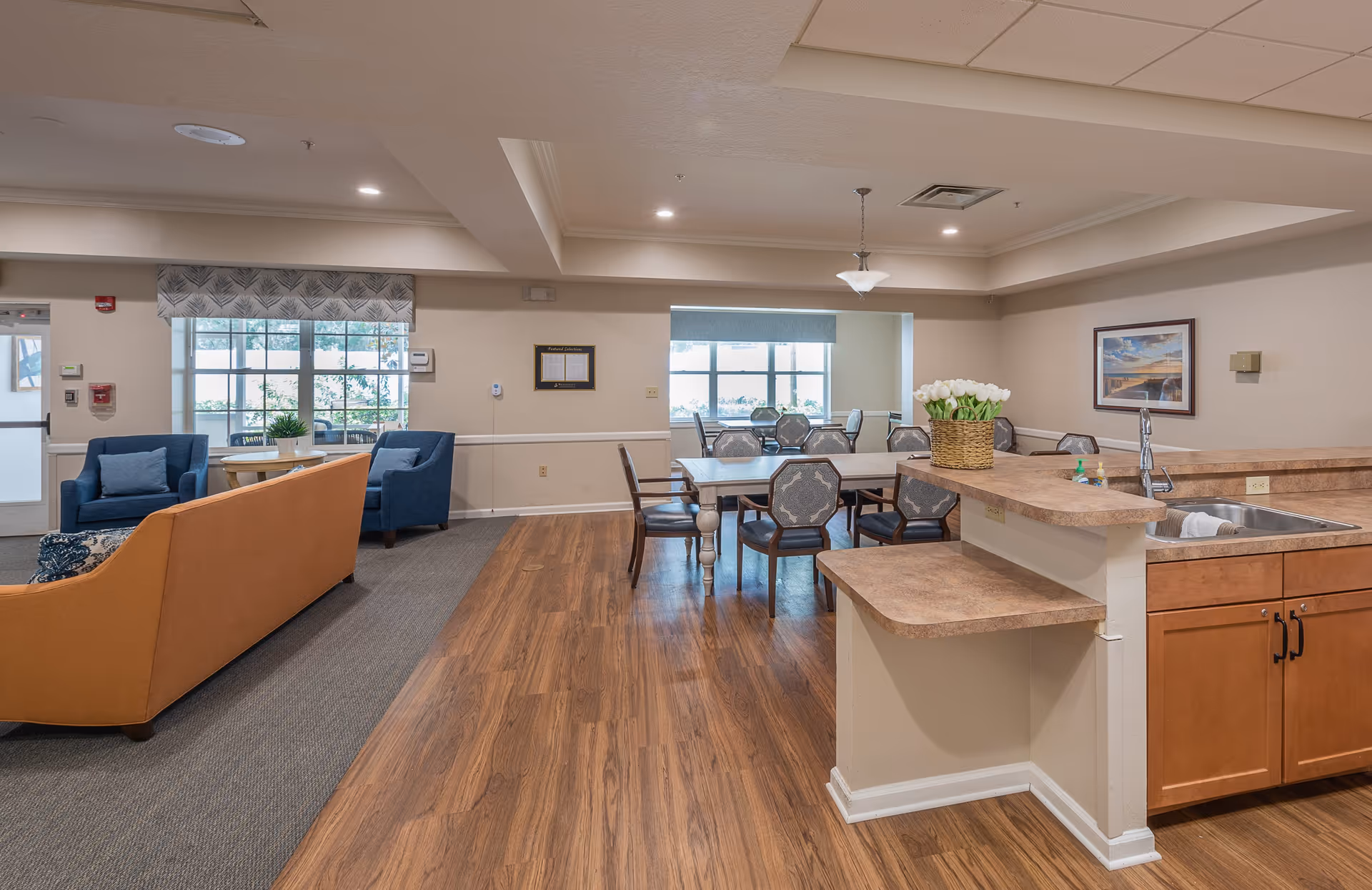 A spacious common area in a senior living facility featuring a kitchen counter with a sink and a basket of white flowers, a dining table with multiple chairs, and a seating area with a brown sofa and two blue armchairs near large windows.