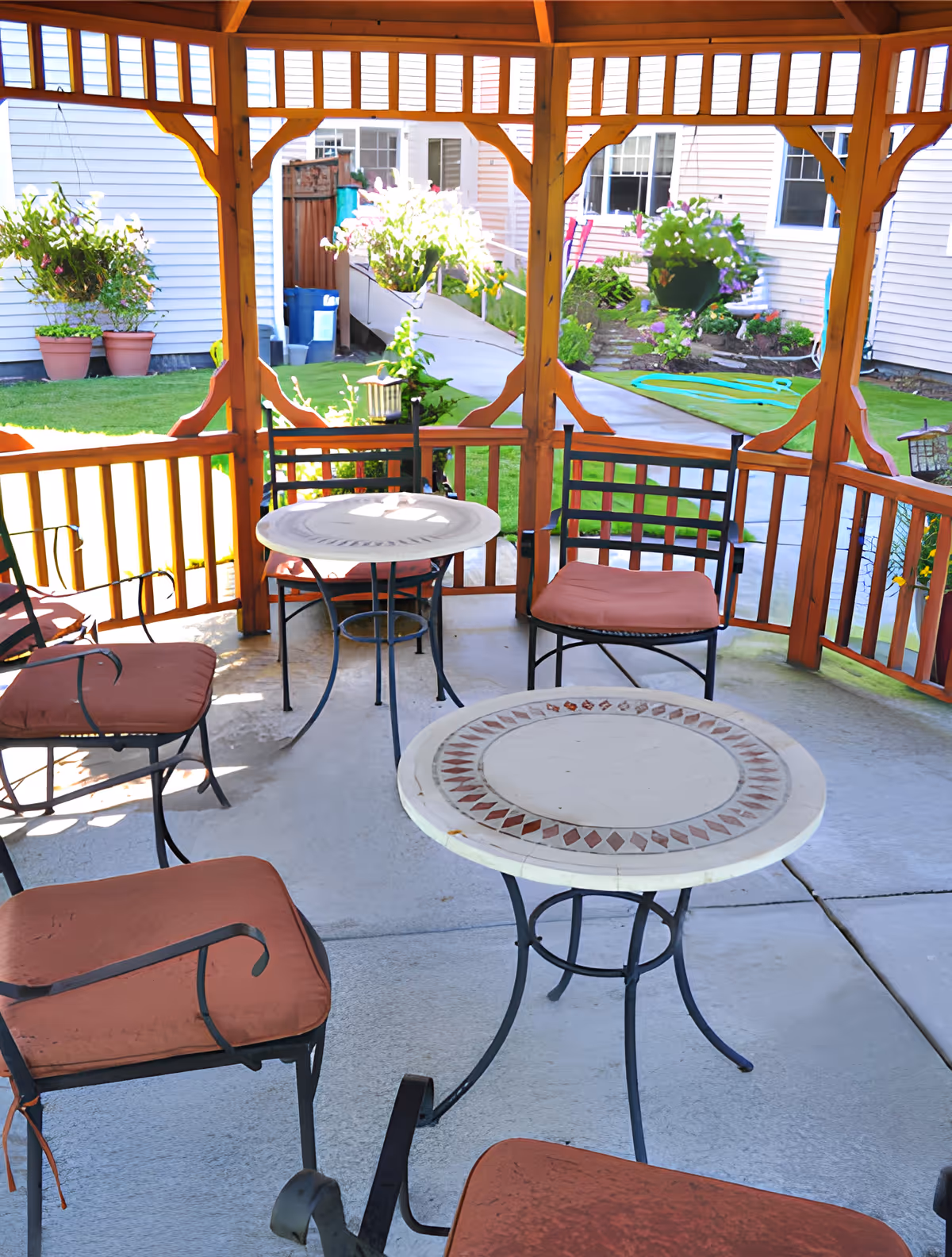 Outdoor covered patio area with a wooden gazebo structure, featuring several cushioned metal chairs and two round mosaic-tiled tables. In the background, there is a garden with green grass, potted plants, flowers, and a concrete walkway leading to a building with white siding.
