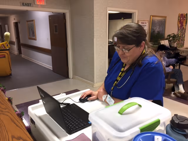 A healthcare worker in blue scrubs with a stethoscope around her neck is working on a laptop placed on a mobile cart in a hallway of a senior care facility. In the background, there is a person sitting on a bench and some wall decorations.