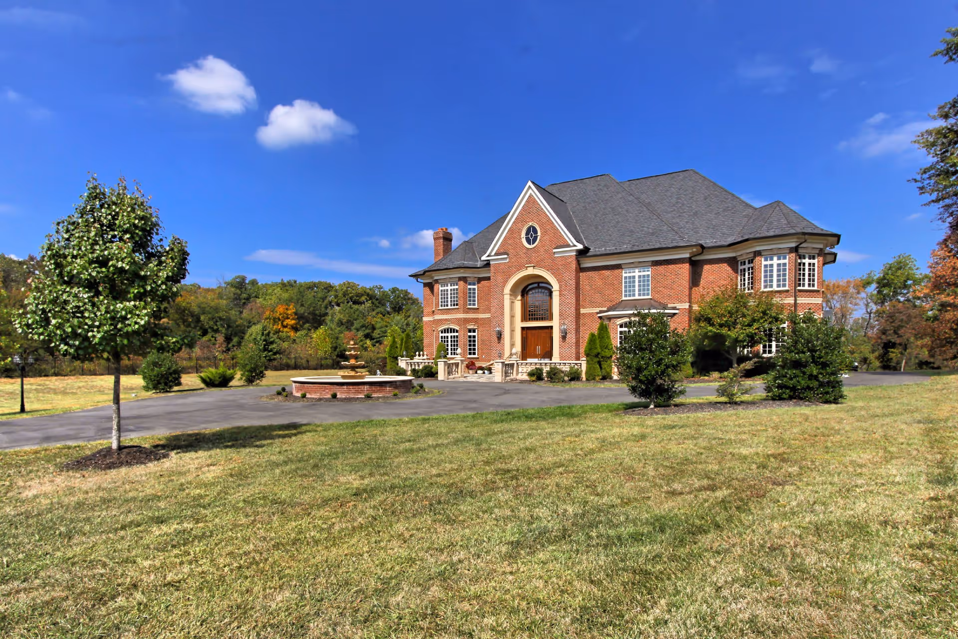 A large, elegant two-story brick building with a gray shingled roof, surrounded by a well-maintained lawn and several small trees. There is a circular driveway with a fountain in front of the building, under a clear blue sky with a few scattered clouds.