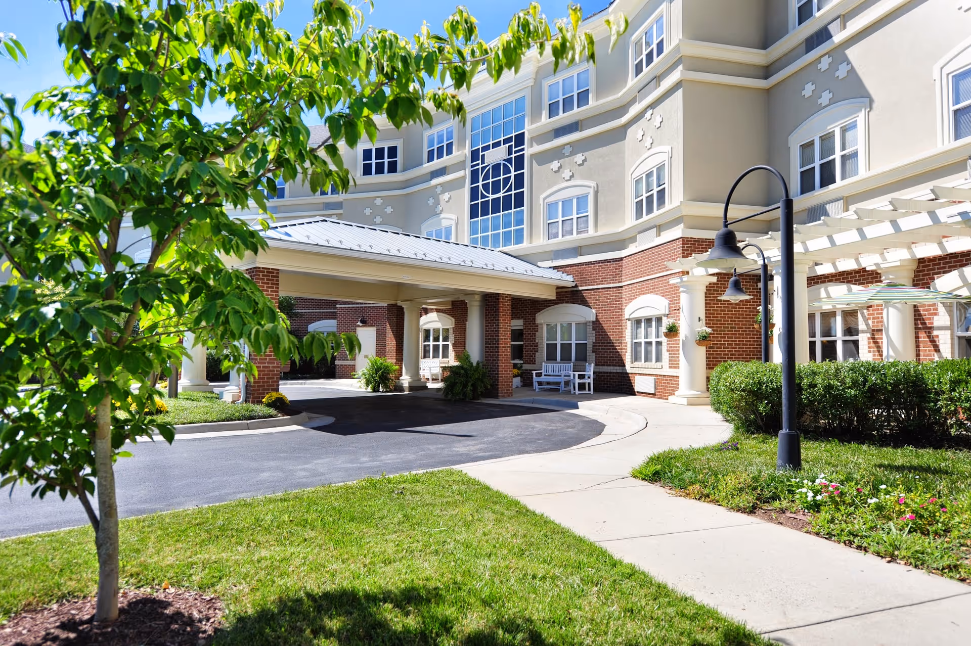 Exterior view of Morningside of Charlottesville senior living facility showing a covered entrance with brick and beige walls, multiple windows, a tree in the foreground, green grass, a sidewalk, and outdoor lighting fixtures under a clear blue sky.