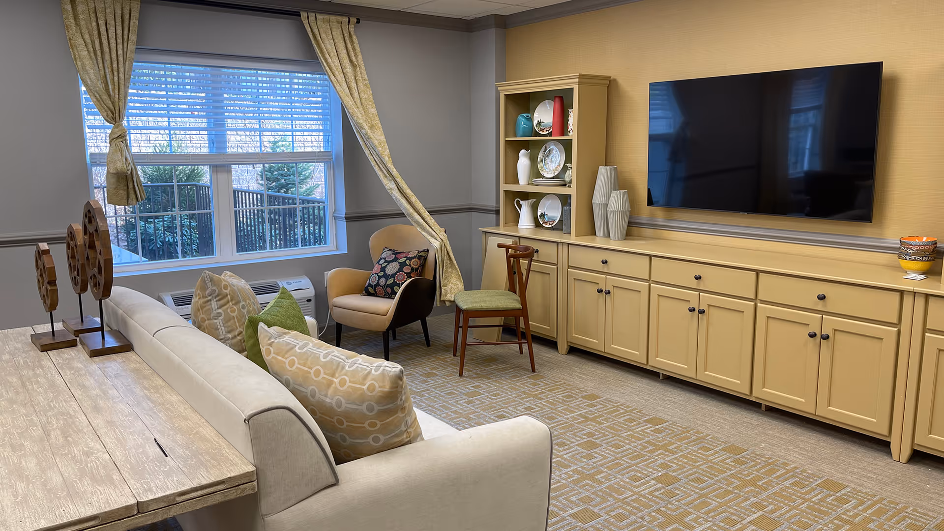 Bright common living room with a sofa, armchairs, built-in cabinets, a wall-mounted TV, and a large window.