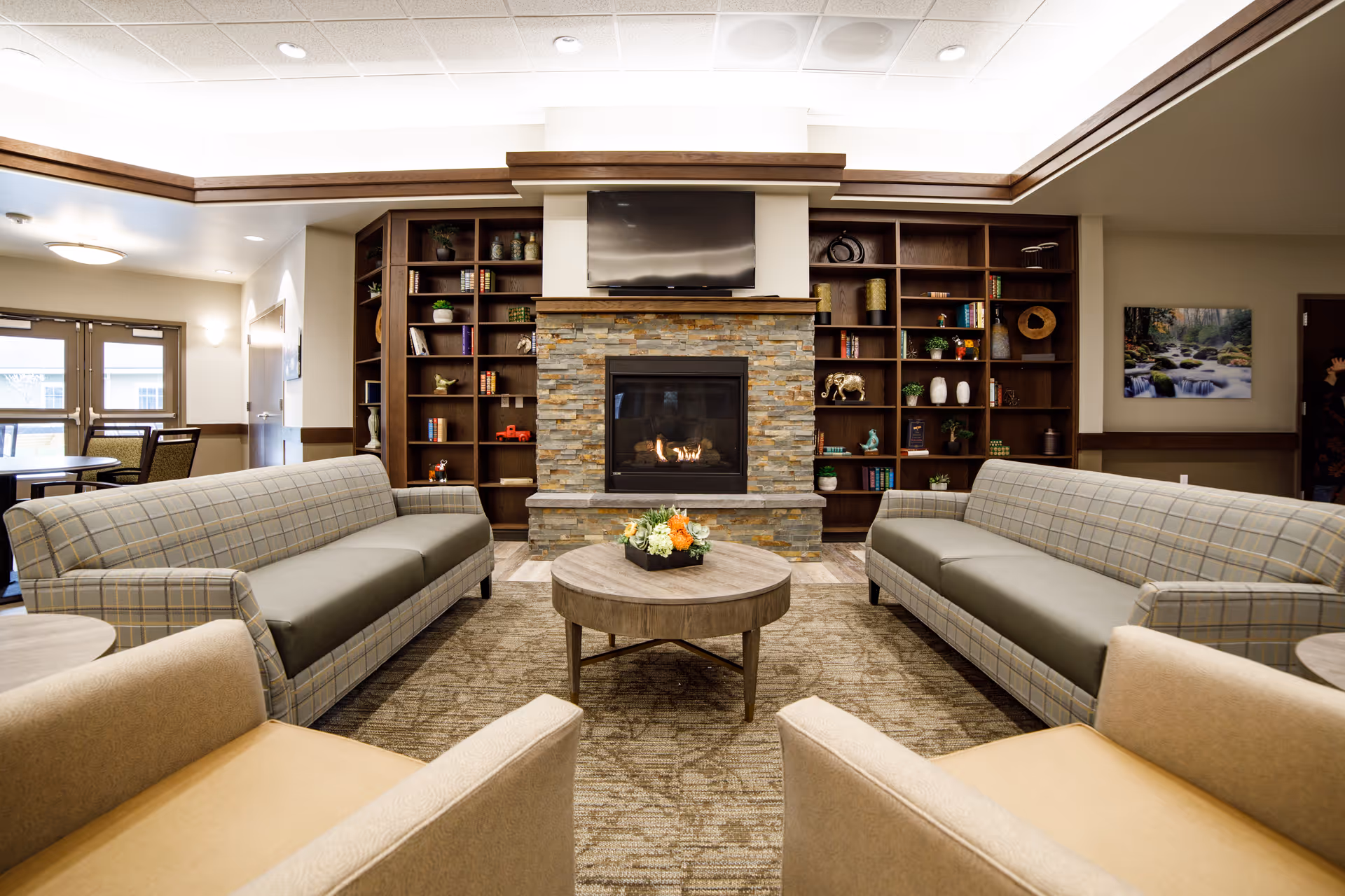 A cozy living room area with two plaid sofas facing each other and two beige armchairs in the foreground. In the center is a round wooden coffee table with a floral arrangement. Behind the seating area is a stone fireplace with a flat-screen TV mounted above it, flanked by built-in wooden bookshelves filled with books and decorative items. The room has a warm, inviting atmosphere with soft lighting and neutral tones.