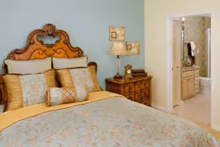 A decorated bedroom with an ornate wooden headboard, a made bed with multiple pillows, a nightstand and lamp, and an open doorway leading to a bathroom.