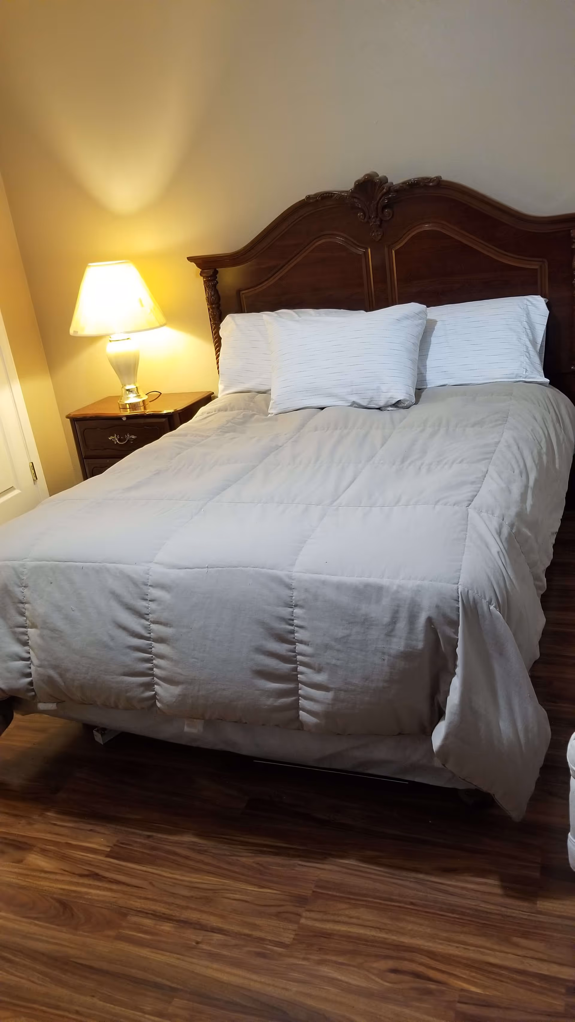 Neatly made bed with a carved wooden headboard, pillows, nightstand and lamp in a small bedroom.