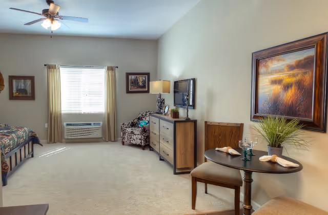 Bright furnished studio-style bedroom and sitting area with a bed, dresser and small dining table.