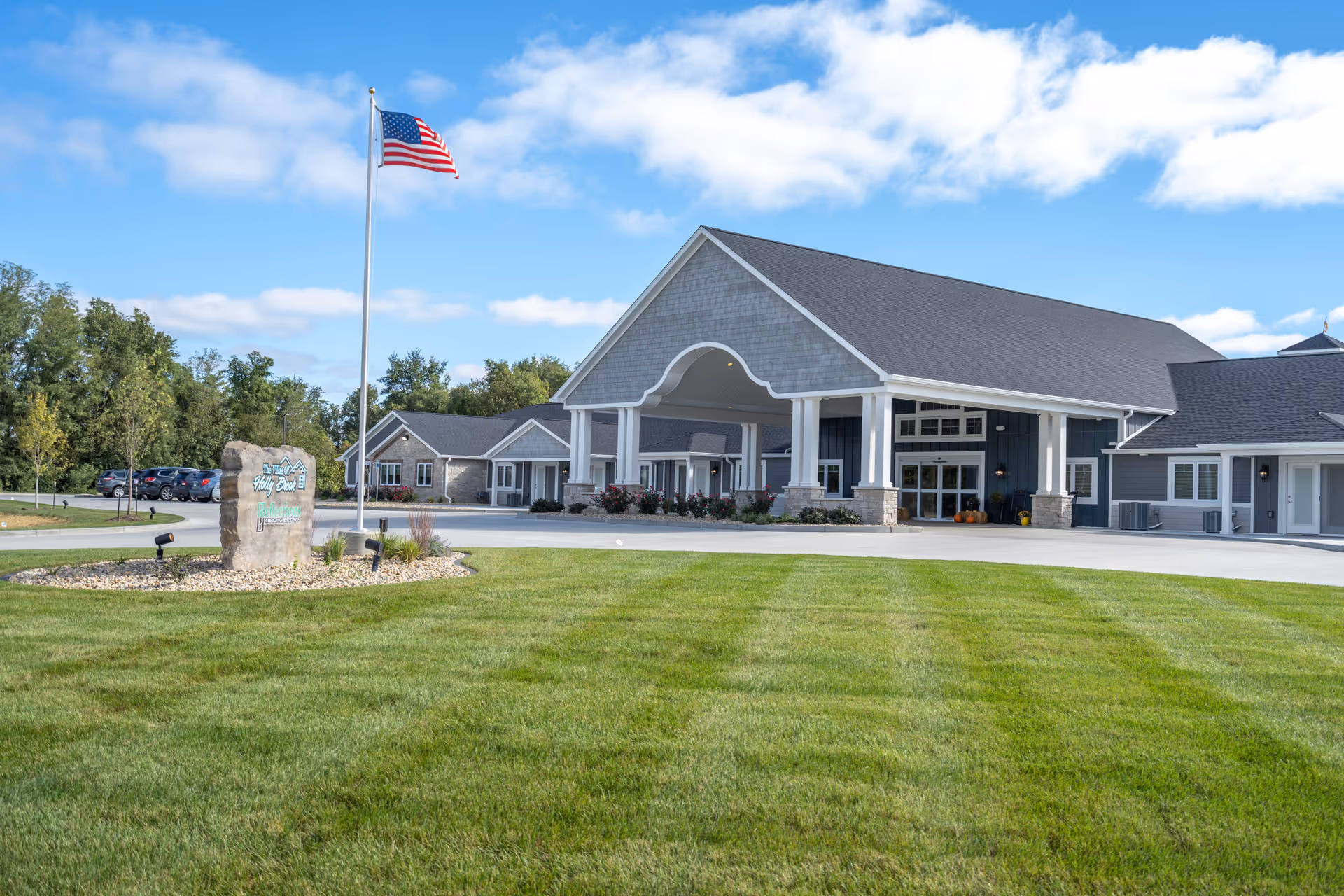 Front exterior of an assisted living building with a large covered entrance, American flag on a flagpole, stone sign, and a manicured lawn.