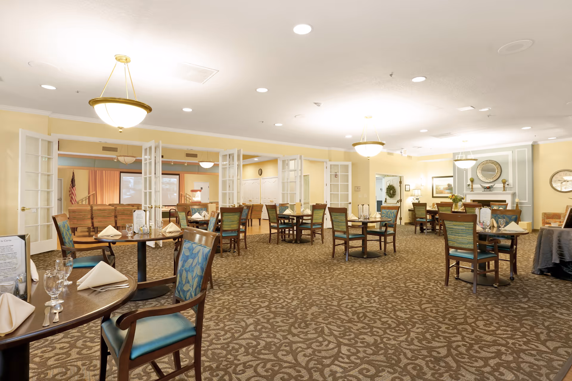 Spacious senior living dining room with multiple set tables and chairs, chandeliers, and open folding doors to an adjoining room.