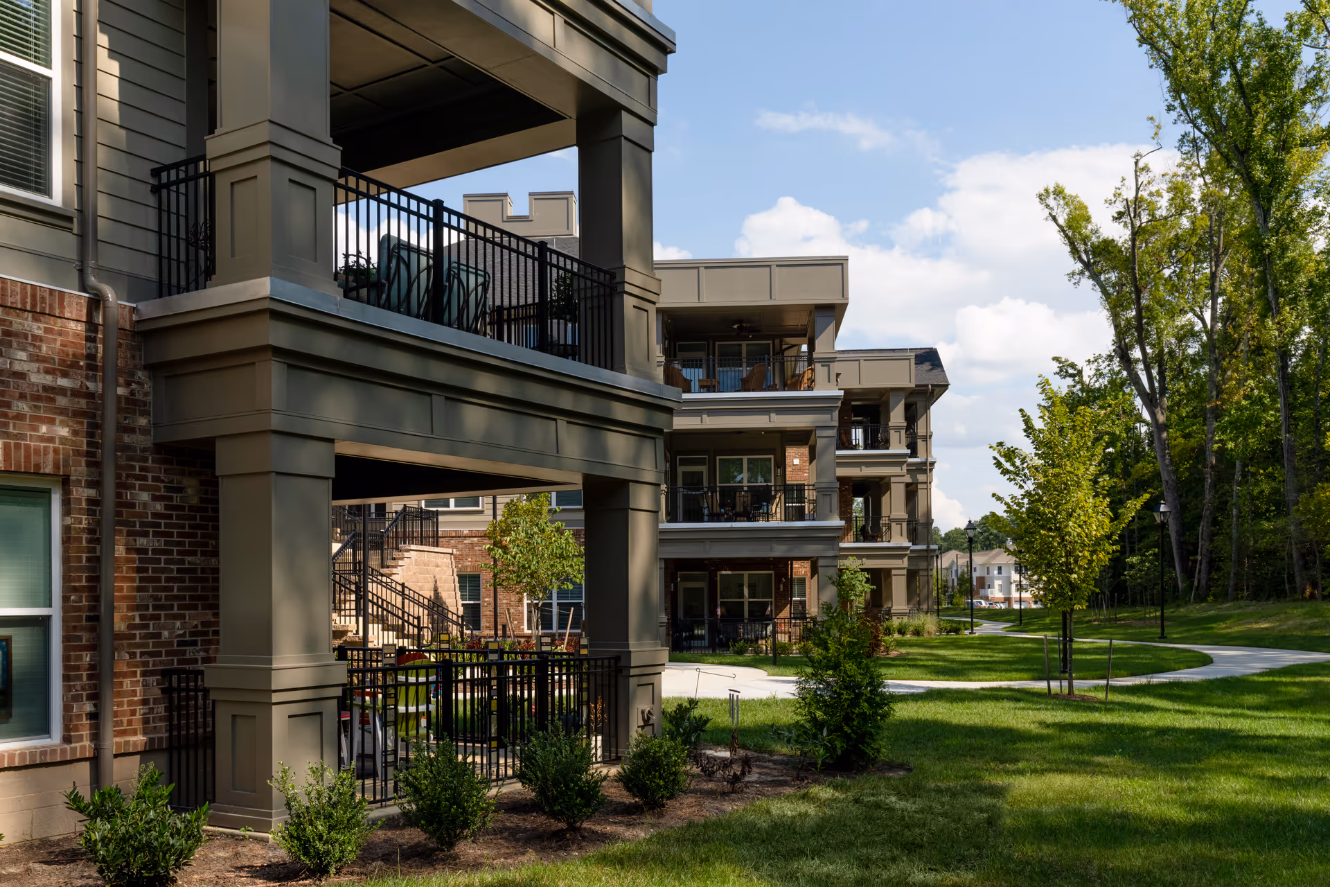 Exterior view of a multi-story residential building with balconies overlooking a well-maintained grassy area with trees and a paved walkway under a partly cloudy sky.