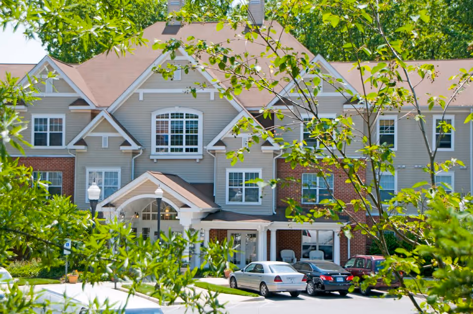 Front entrance of a multi-story senior living building with a covered entry, parked cars, and trees in the foreground.