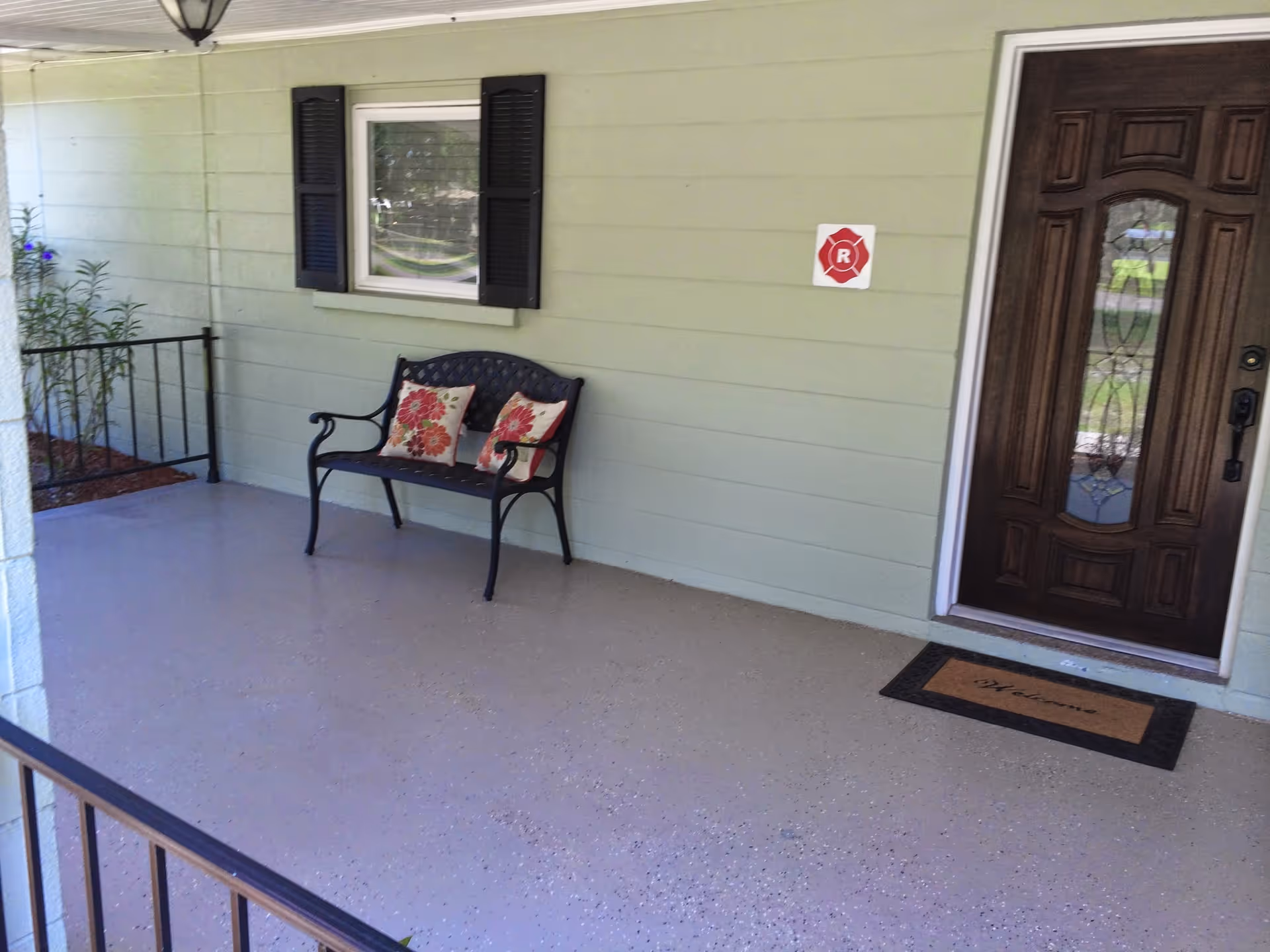 A covered porch area with a black metal bench that has two floral cushions. There is a window with black shutters on the light green exterior wall and a dark wooden front door with decorative glass. A welcome mat is placed in front of the door, and a small plant is visible to the left side near a black railing.