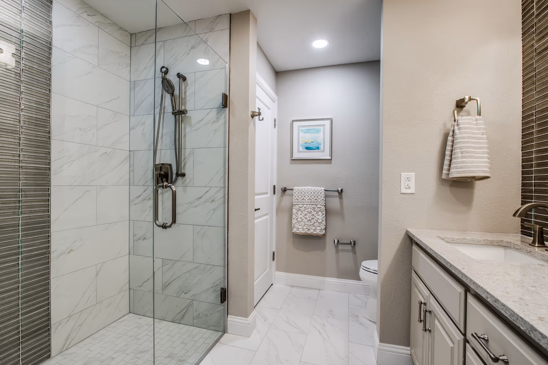 Modern bathroom with a glass-enclosed shower featuring marble-like tiles, a white toilet, a vanity with a sink and granite countertop, a towel hanging on a ring, and a framed abstract artwork on the wall.