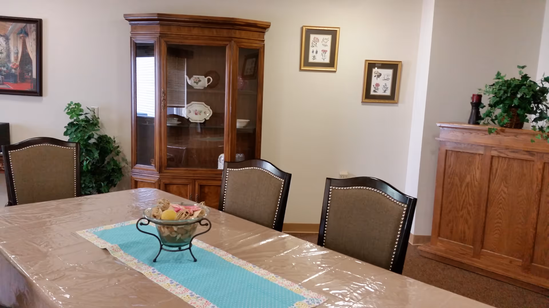 Interior view of a dining room with a table covered by a plastic tablecloth and a decorative runner. The table has a centerpiece bowl with decorative items. There are upholstered chairs around the table. In the background, there is a wooden china cabinet with dishes inside, framed botanical prints on the wall, a wooden sideboard with a plant and candle on top, and a potted plant in the corner.