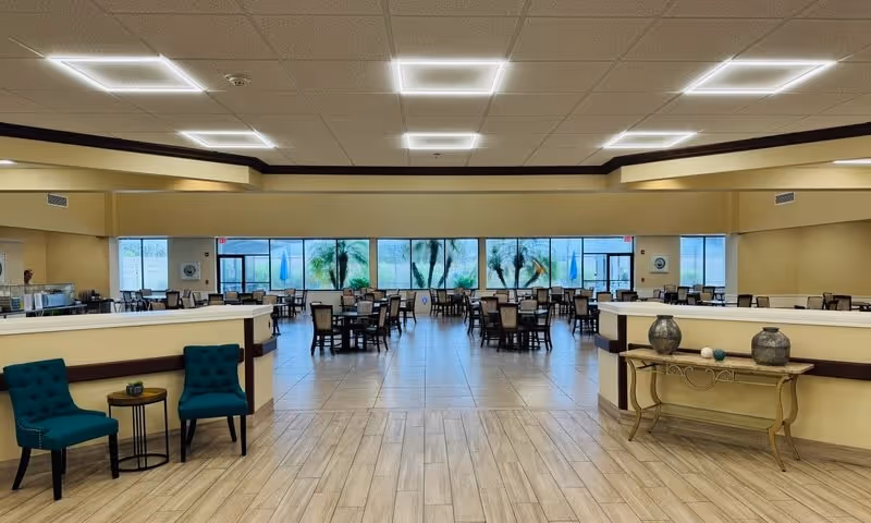 Spacious dining room with rows of tables and chairs, teal accent chairs and a decorative console in the foreground, and large windows along the back wall.