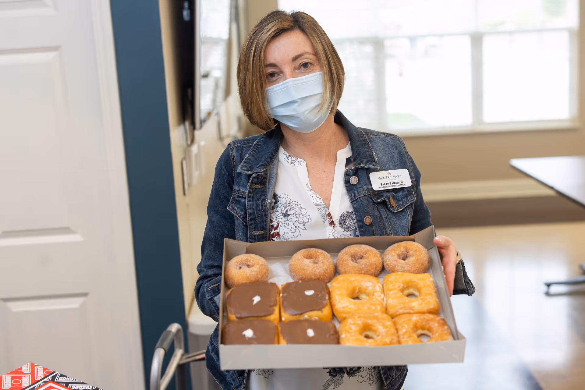 A woman wearing a face mask and a denim jacket holds a box of assorted donuts, including glazed, chocolate-topped, and sugar-coated varieties, inside a well-lit room with large windows.