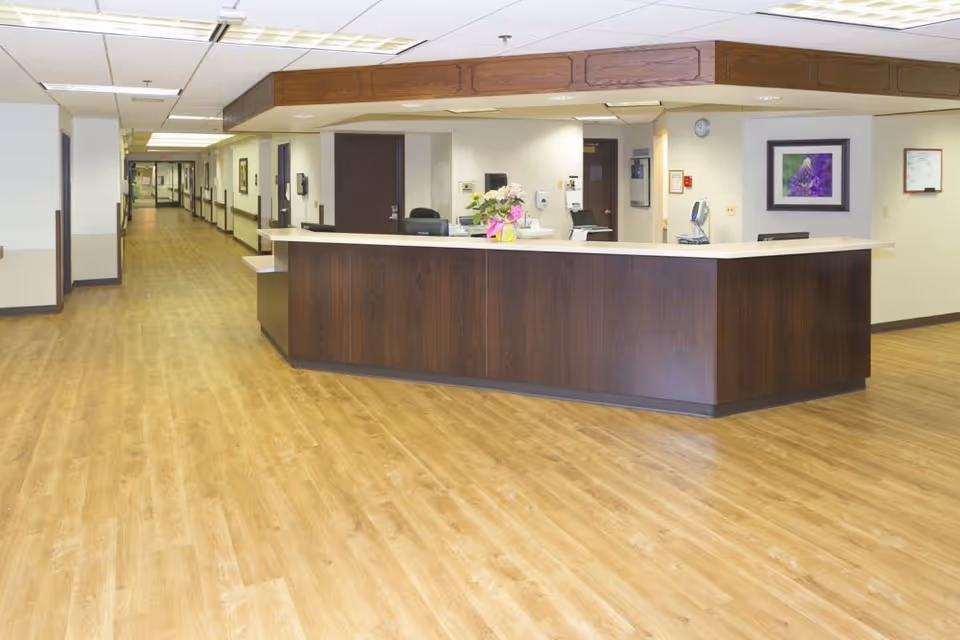 Reception desk area in a senior living facility with wood flooring, a long hallway with doors on either side, and a flower arrangement on the counter. The walls are light-colored with framed artwork and a clock visible.