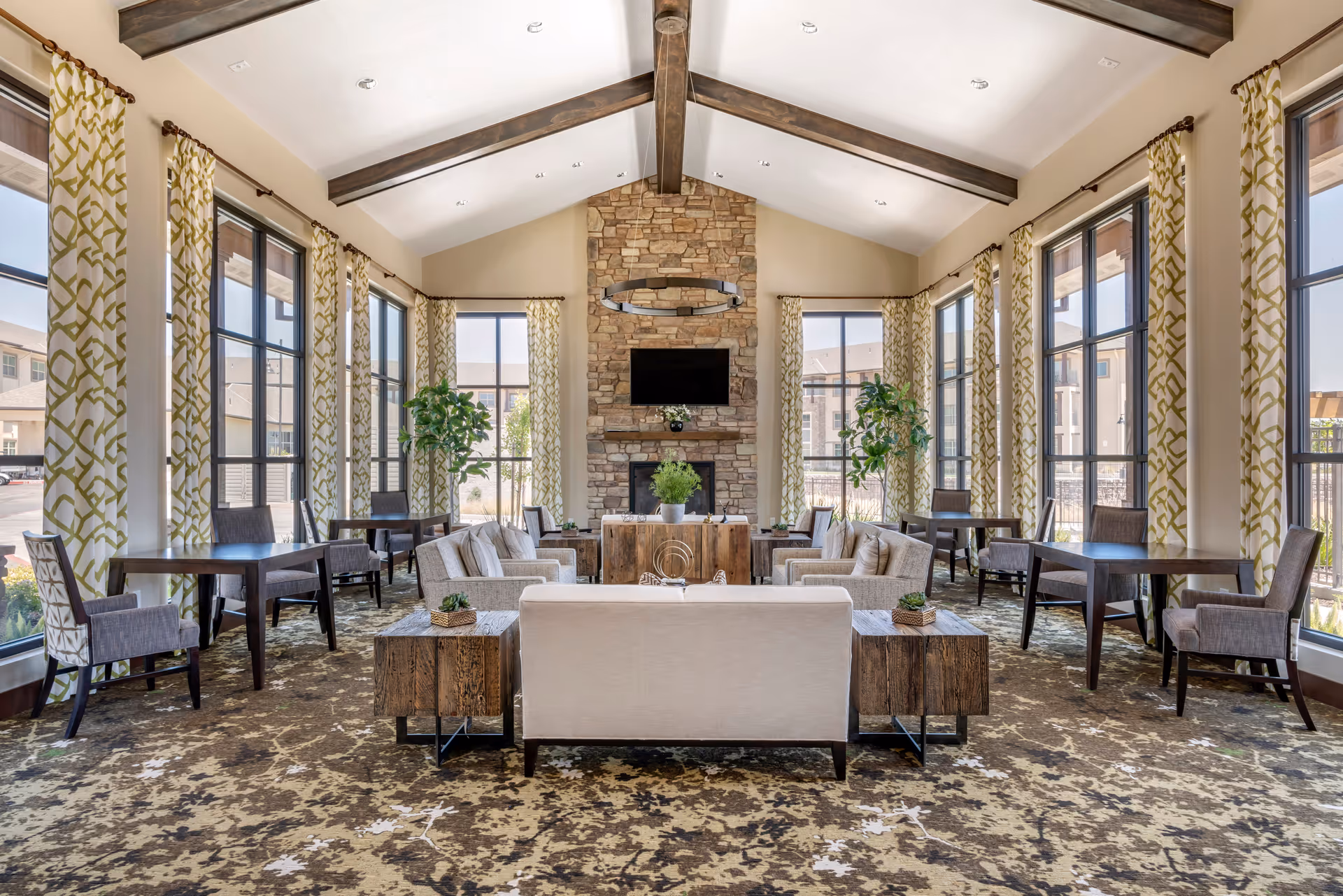 Spacious senior living lounge with high beamed ceiling, stone fireplace, sofas, tables, and large windows.