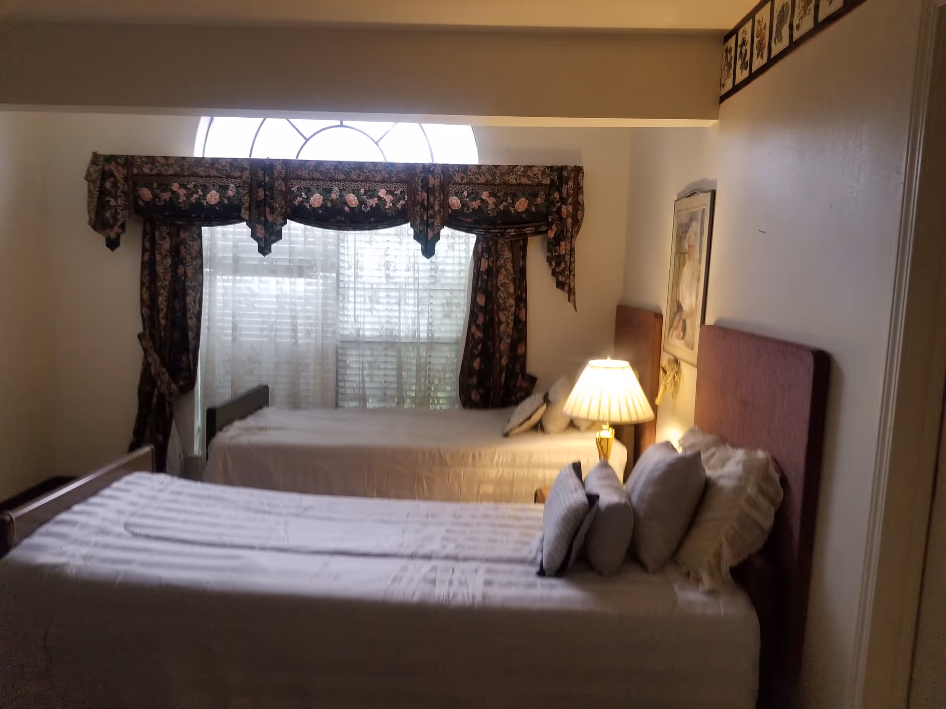 A bedroom with two single beds placed parallel to each other. Each bed has white bedding and multiple pillows. A table lamp with a beige shade is on a nightstand between the beds. The room has a large window with floral-patterned curtains and sheer white blinds. A framed picture hangs on the wall above one of the beds.