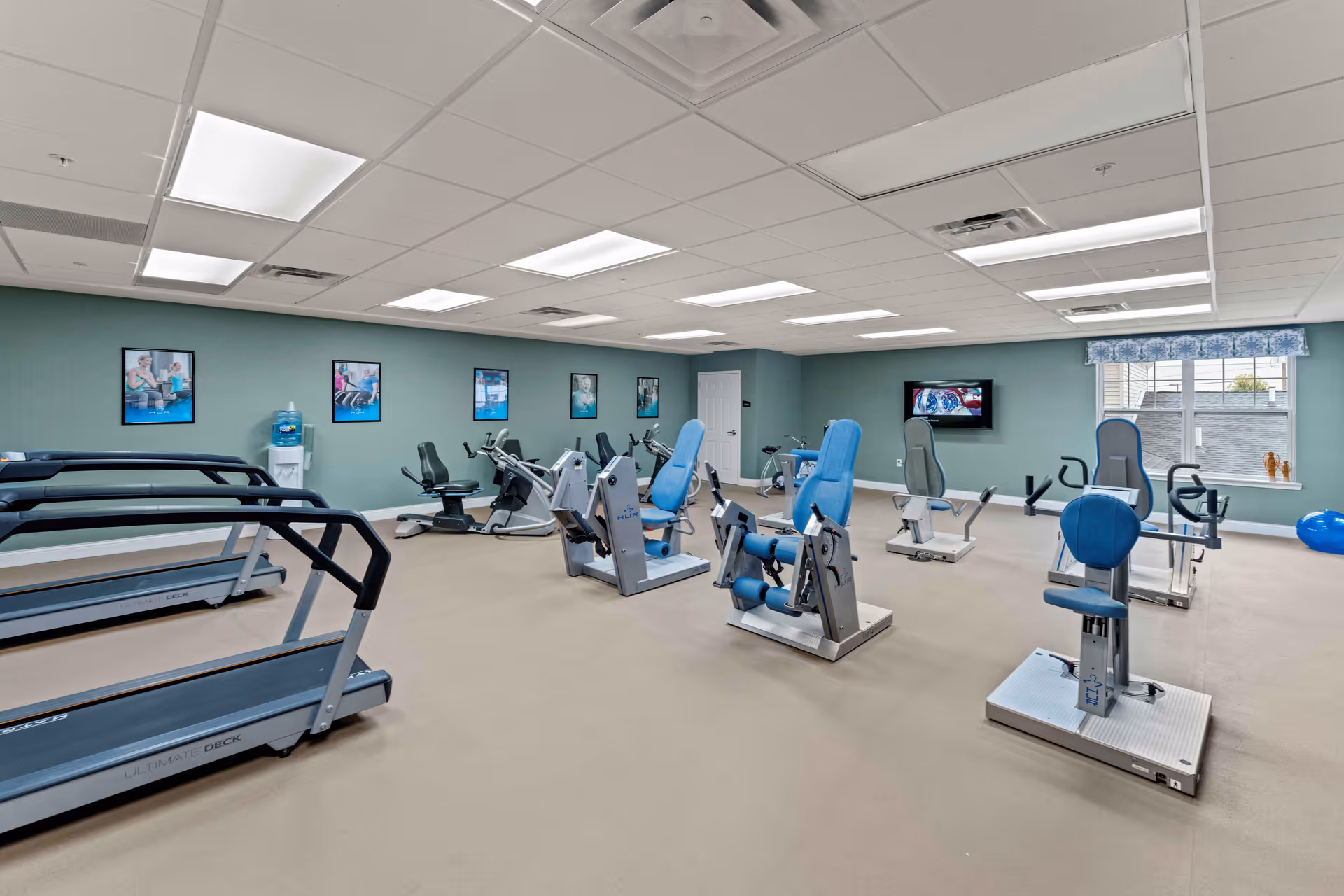 A spacious fitness room with multiple exercise machines including treadmills, stationary bikes, and strength training equipment. The room has light green walls adorned with framed pictures, a water cooler, a wall-mounted TV, and a window with a blue valance letting in natural light.