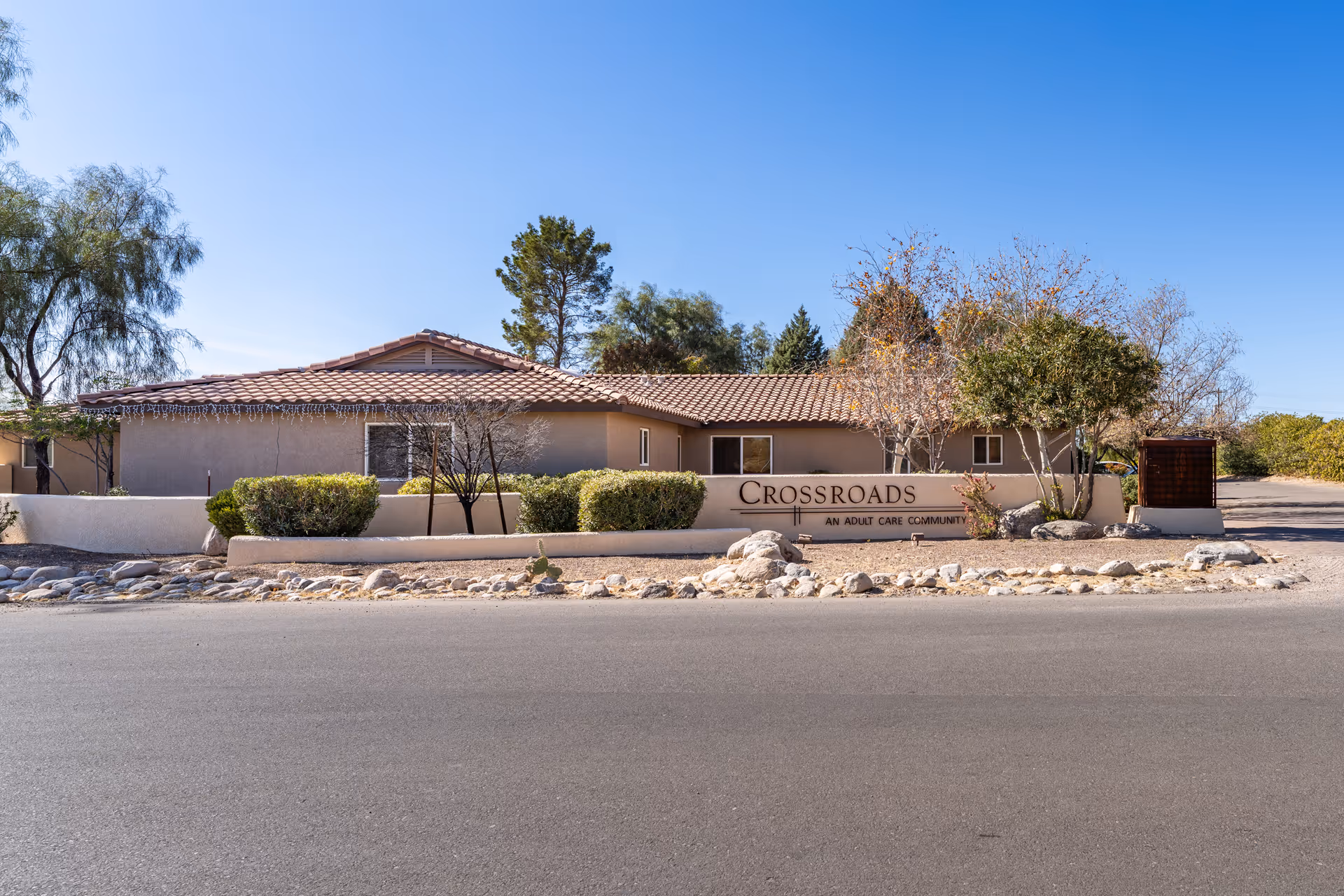 Single-story stucco assisted living building with a tiled roof and a 'Crossroads' sign in front, surrounded by shrubs and desert landscaping.