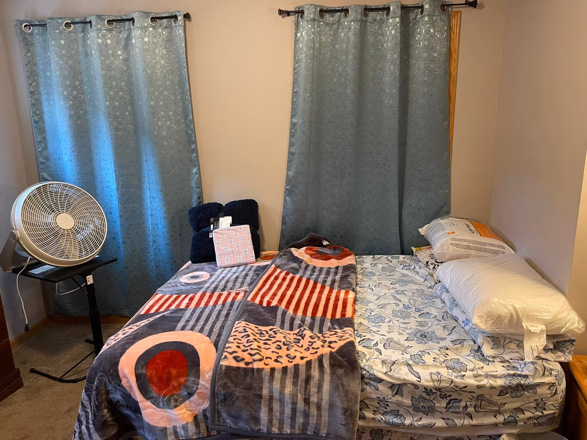 A small bedroom with two twin beds pushed together, one bed covered with a colorful patterned blanket and the other with floral sheets and pillows stacked on top. Behind the beds are two windows with blue curtains. A white fan is placed on a small black stand to the left of the beds.