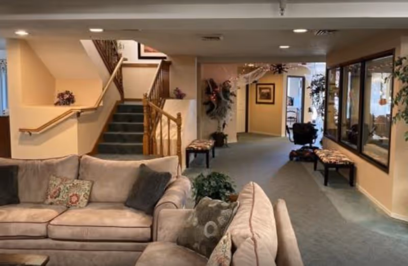 Interior view of a senior living facility showing a cozy seating area with beige sofas and patterned cushions in the foreground. Behind the seating area is a carpeted hallway with benches along the wall, decorative plants, and framed artwork. A staircase with wooden railings is visible on the left side, leading to an upper floor. The space is softly lit with recessed ceiling lights.