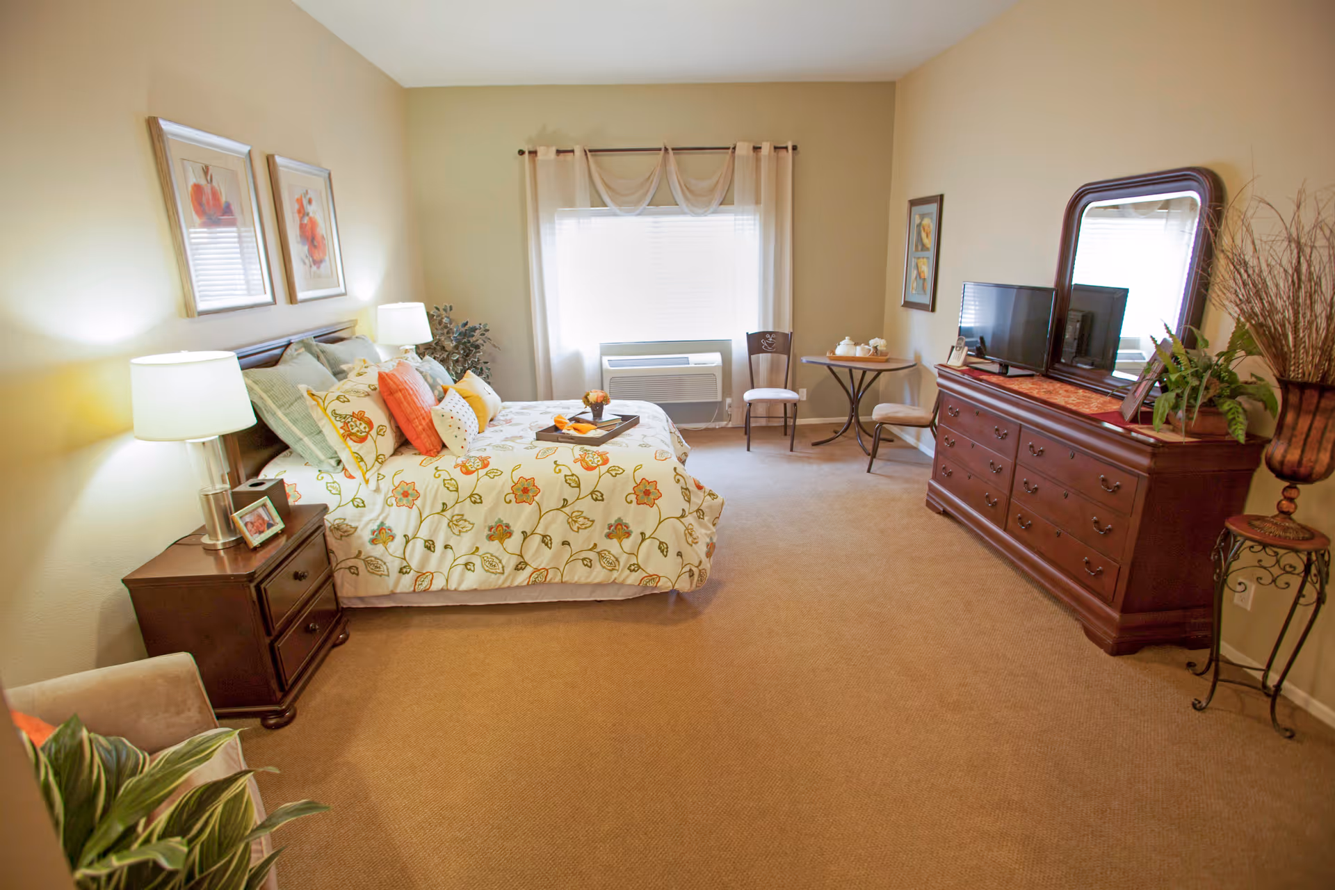 Well-lit furnished bedroom with a bed, nightstands, a dresser with mirror and TV, and a small seating area.