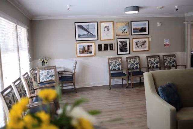 A well-lit common area with wooden flooring and light gray walls decorated with framed pictures. There are several chairs with floral patterned cushions arranged along the walls and a small table with two chairs near a window with blinds. A beige sofa with a blue cushion is partially visible in the foreground, and a vase with yellow flowers is blurred in the front.