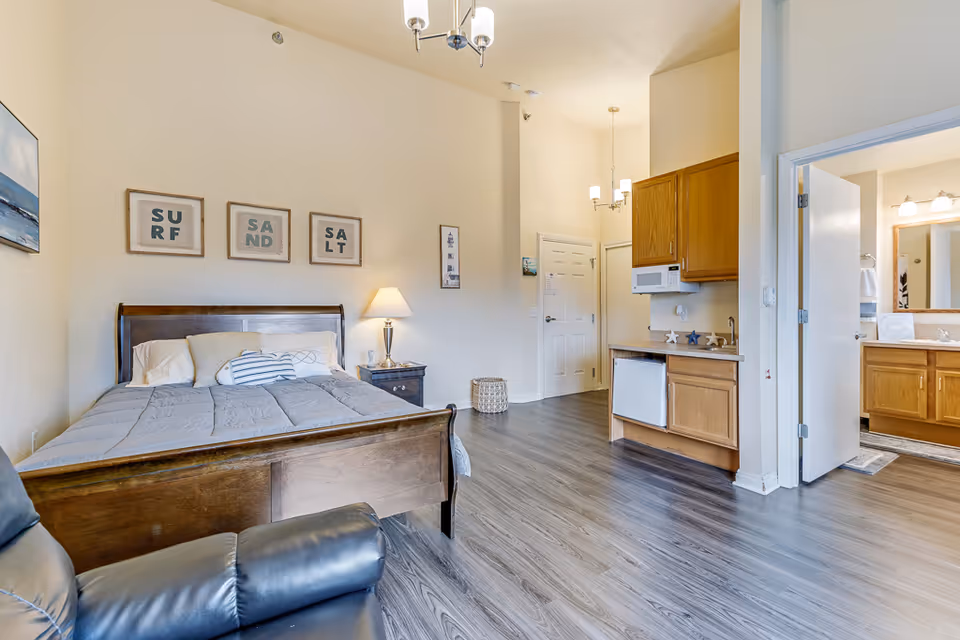 A senior living facility room featuring a wooden bed with gray bedding and pillows, a nightstand with a lamp, a black leather recliner chair, and wall art with the words 'SURF', 'SAND', and 'SALT'. The room has wood flooring, a small kitchenette area with a microwave, mini fridge, and sink, and an open door leading to a bathroom with a large mirror and wooden cabinets.