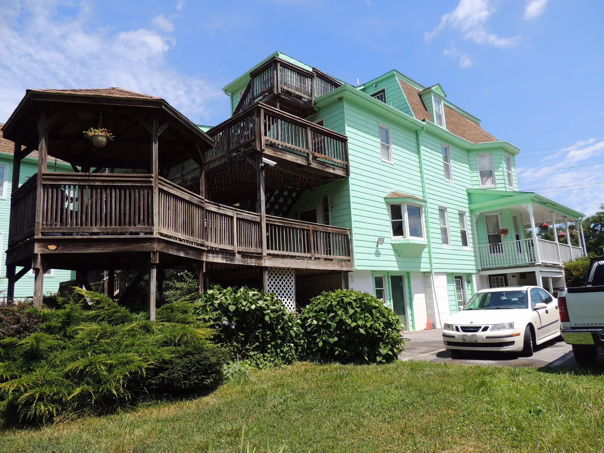 Exterior view of a light green multi-story assisted living facility with wooden balconies and a gazebo-like structure. There are bushes and grass in the foreground and two parked cars near the building under a clear blue sky.