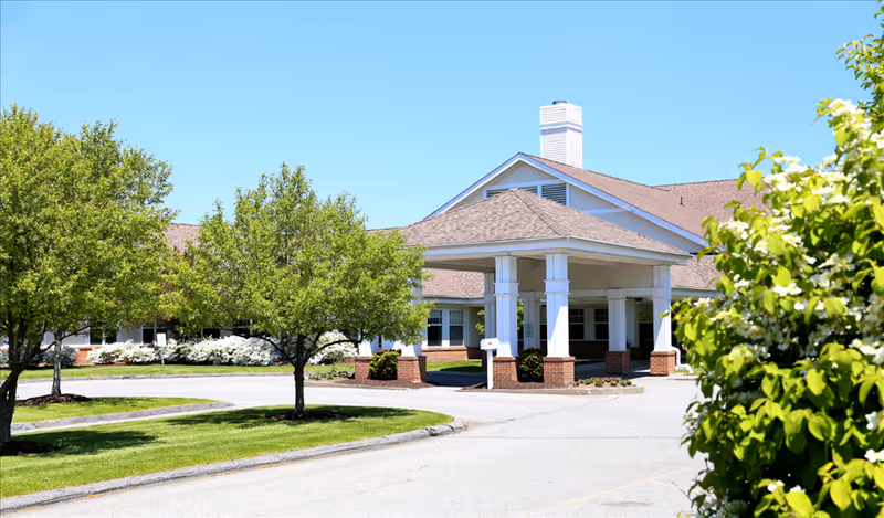 Exterior view of Putnam Ridge facility showing the entrance with a covered drop-off area supported by white columns with brick bases, surrounded by green trees and shrubs under a clear blue sky.
