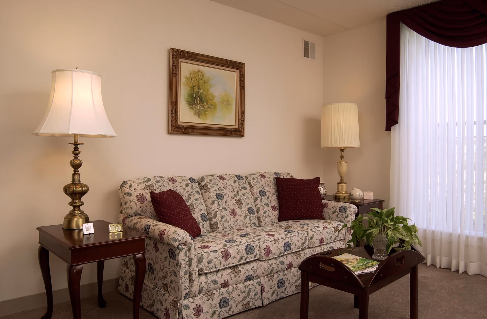 A cozy living room area with a floral patterned sofa adorned with two burgundy pillows. On either side of the sofa are wooden side tables, each with a brass lamp. A framed painting of a tree by a lake hangs on the wall above the sofa. A wooden coffee table in front of the sofa holds a green plant and some magazines. Light filters in through a window with sheer white curtains and burgundy valances.