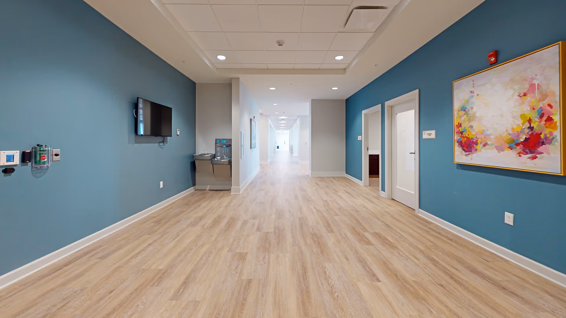 A long, wide hallway in an assisted living facility with light wood flooring and teal blue walls. On the left wall, there is a mounted flat-screen TV and a water fountain below it. On the right wall, there is a colorful abstract painting and two white doors. The hallway is well-lit with recessed ceiling lights and extends into the distance.