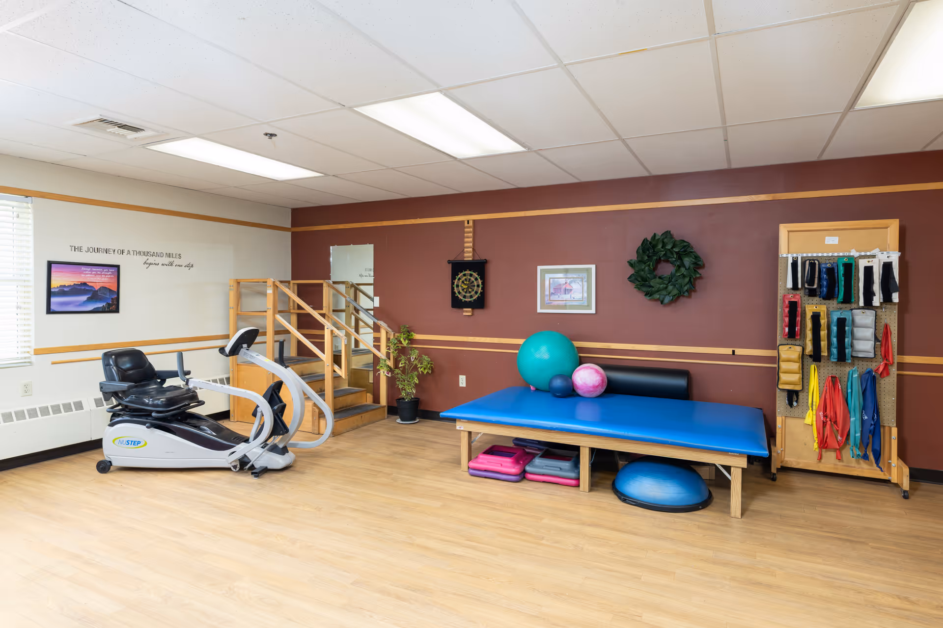 Therapy/exercise room with a recumbent stepper, a blue treatment table, exercise balls, and a rack of resistance bands and equipment.