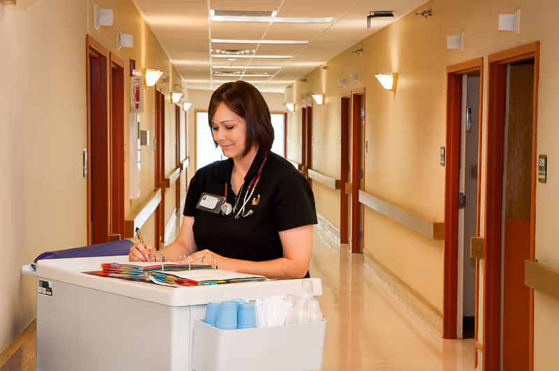 A healthcare worker with a stethoscope around her neck is writing in a binder on a mobile workstation cart in a well-lit hallway of a senior care facility.