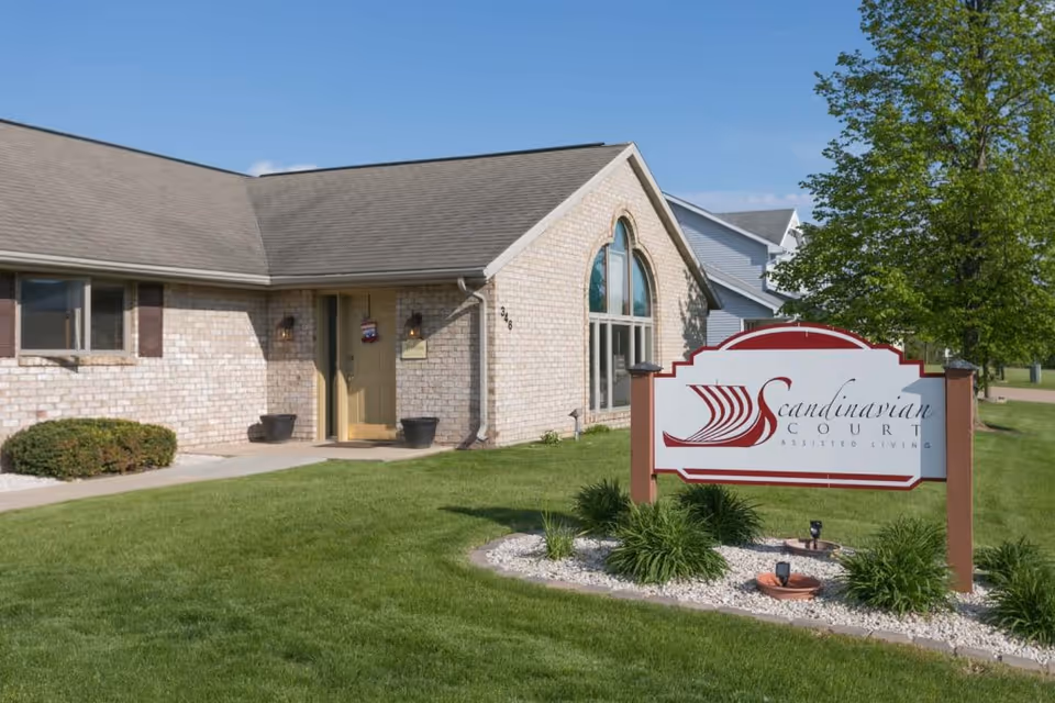 Exterior view of a single-story brick building with a gable roof, a front door with two wall-mounted lights, and a large arched window. In front of the building is a landscaped lawn with green grass, bushes, and a sign that reads 'Scandinavian Court Assisted Living'. A tree is visible on the right side under a clear blue sky.