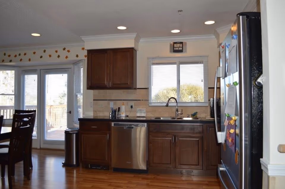 A kitchen with wooden cabinets, a stainless steel dishwasher, a double sink under a window with blinds, and a stainless steel refrigerator with magnets and papers attached. To the left, there is a dining area with a wooden table and chairs, and glass doors leading to an outdoor deck. The walls have a decorative border with autumn leaves near the ceiling.