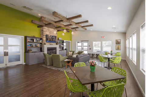 A bright and modern common area in a senior living facility featuring a seating area with green and gray chairs around a stone fireplace, wooden ceiling beams, and a TV mounted above the fireplace. There are several tables with green chairs near large windows letting in natural light. The walls are painted white and green, and the floor is wood.