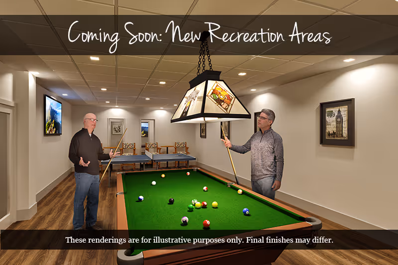 Two men stand by a pool table in a recreation room with a hanging lamp and a ping-pong table in the background.